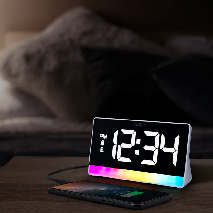 Lifestyle image of the 617-149 Digital Alarm with Color Changing Glow Light shown on a nightstand with a bed in the background in a darkened bedroom. The alarm clock features a white case, a color-changing bar as the base, and a black display with white digits.