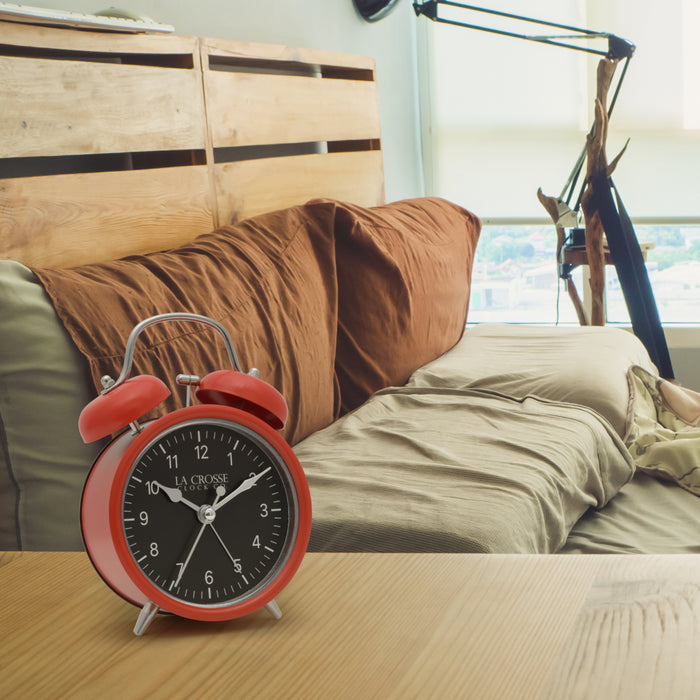 Lifestyle image of the 617-3314 Metal Twin Bell Alarm Clock – Red, sitting on a nightstand with a bedroom scene in the background. The clock features a black dial with white numbers, silver hands and accents, in a red metal case