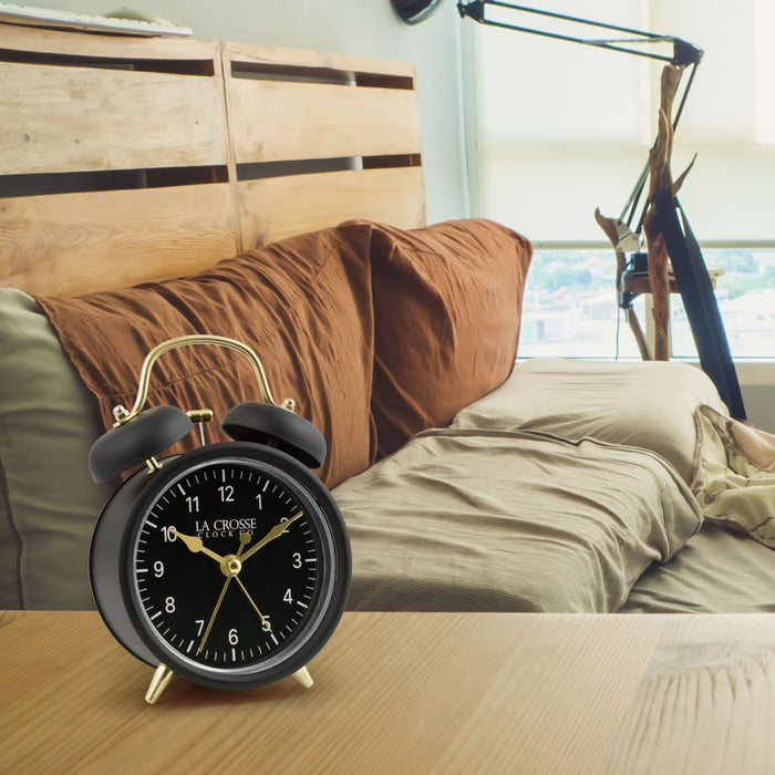 Lifestyle image of the 617-3314BG Metal Twin Bell Alarm Clock – Black, sitting on a nightstand with a bedroom scene in the background. The clock features a black dial with white numbers, gold hands and accents, in a black metal case.