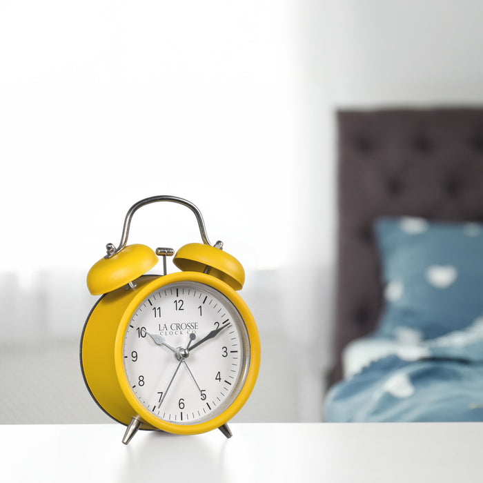 Lifestyle image of the 617-3314MY Metal Twin Bell Alarm Clock – Mustard Yellow, sitting on a nightstand with a bedroom scene in the background. The clock features a white dial with black numbers, silver hands and accents, in a mustard yellow metal case.