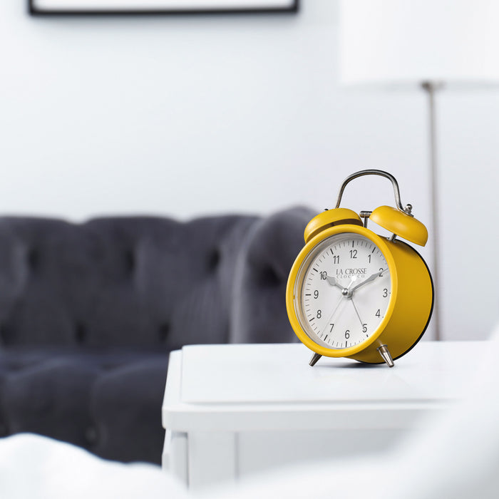 Lifestyle image of the 617-3314MY Metal Twin Bell Alarm Clock – Mustard Yellow, sitting on a white side table with a dark grey couch in the background. The clock features a white dial with black numbers, silver hands and accents, in a mustard yellow metal case.