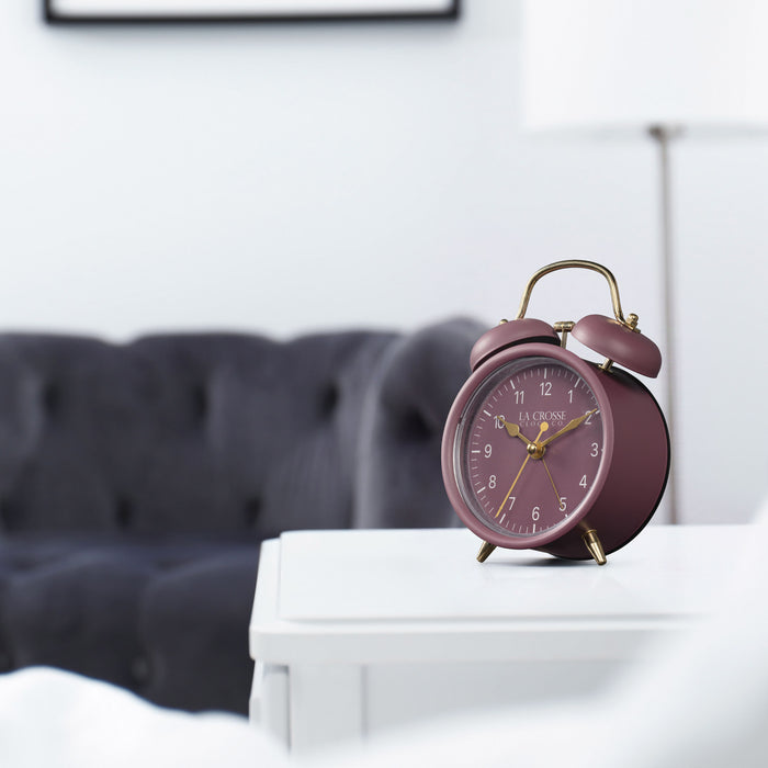 Lifestyle image of the 617-3314RR Metal Twin Bell Alarm Clock – Ruby Red, sitting on a white side table with a dark grey couch in the background. The clock features a ruby red dial with white numbers, gold hands and accents, in a ruby red metal case.