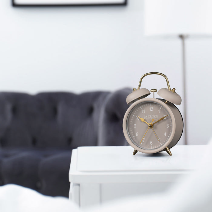 Lifestyle image of the 617-3314TP Metal Twin Bell Alarm Clock – Taupe, sitting on a nightstand with a bedroom scene in the background. The clock features a taupe dial with white numbers, gold hands and accents, in a taupe metal case.