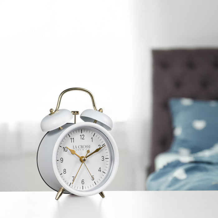 Lifestyle image of the 617-3314W Metal Twin Bell Alarm Clock – White, sitting on a nightstand with a bedroom scene in the background. The clock features a white dial with black numbers, gold hands and accents, in a white metal case.