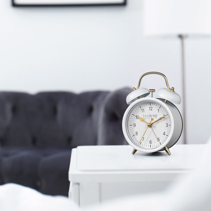 Lifestyle image of the 617-3314W Metal Twin Bell Alarm Clock – White, sitting on a white side table with a dark grey couch in the background. The clock features a white dial with black numbers, gold hands and accents, in a white metal case.