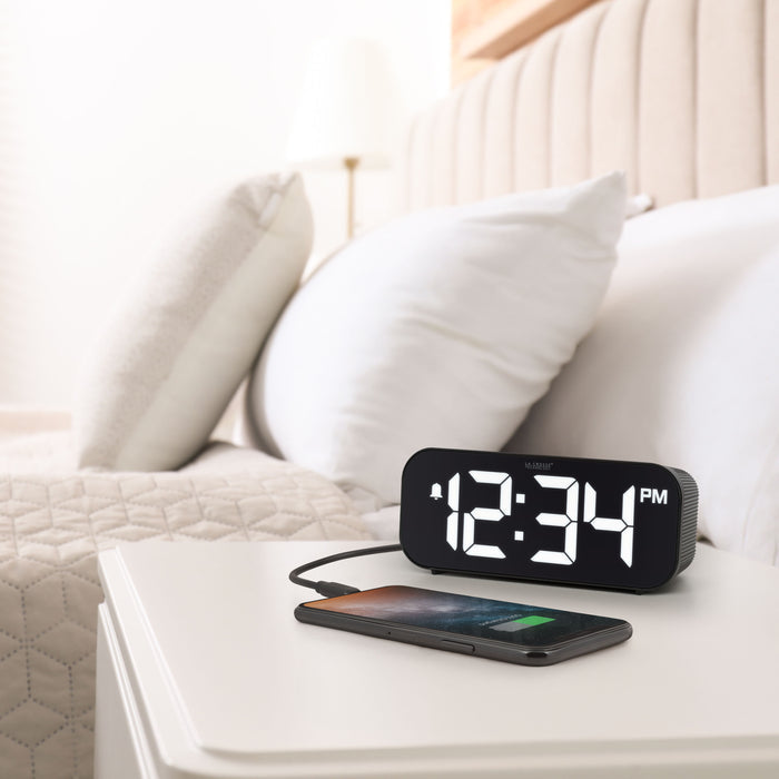Lifestyle image of the 617-714 Insta-Set Digital LED Alarm Clock with Bluetooth® Technology - Black sitting on a white nightstand with a bed in the background. The alarm clock is shown with a smartphone being charged. The alarm clock features large white digits, a PM indicator, and an alarm icon in a black case.