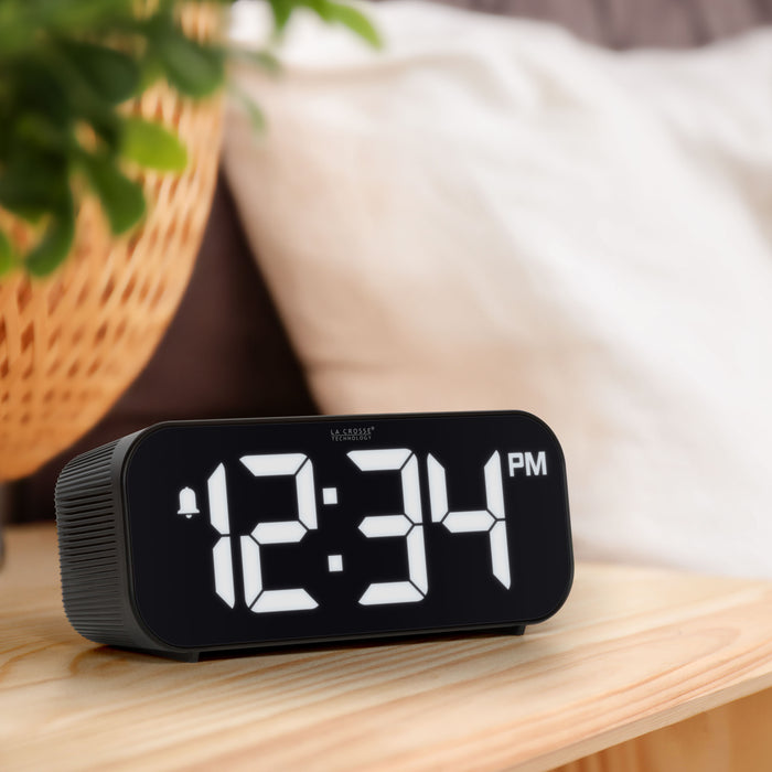 Lifestyle image of the 617-714 Insta-Set Digital LED Alarm Clock with Bluetooth® Technology in Black sitting on a wood nightstand. The alarm clock features large white digits, a PM indicator, and an alarm icon in a black case.