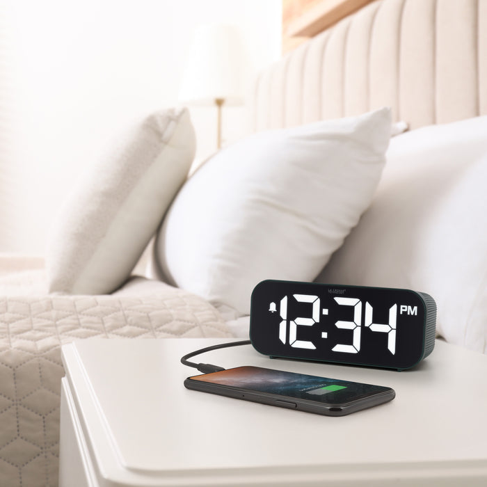 Lifestyle image of the 617-714G Insta-Set Digital LED Alarm Clock with Bluetooth® Technology - Green sitting on a white nightstand with a bed in the background. The alarm clock is shown with a smartphone being charged. The alarm clock features large white digits, a PM indicator, and an alarm icon in a green case.