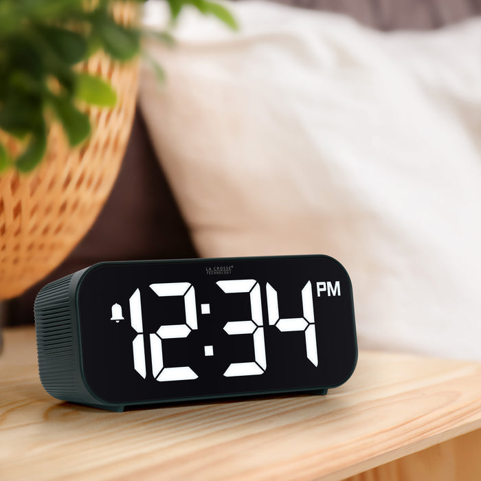 Lifestyle image of the 617-714G Insta-Set Digital LED Alarm Clock with Bluetooth® Technology - Green sitting on a wood nightstand. The alarm clock features large white digits, a PM indicator, and an alarm icon in a green case.