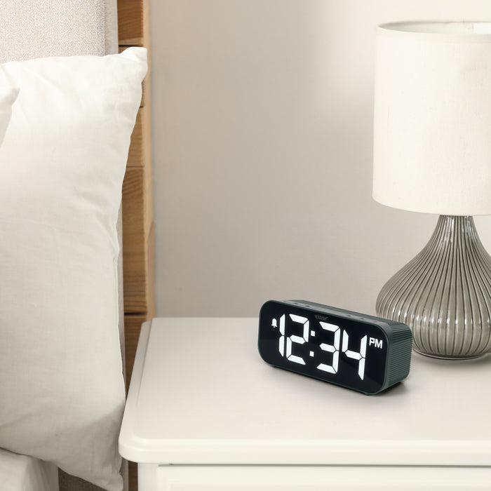 Lifestyle image of the 617-714G Insta-Set Digital LED Alarm Clock with Bluetooth® Technology - Green sitting on a white nightstand with a silver base lamp. The alarm clock features large white digits, a PM indicator, and an alarm icon in a green case.