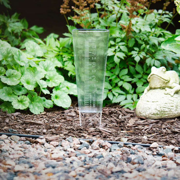 704-5331 USA-Made Clear Measure Rain Gauge lifestyle in a plant bed with bark colored rocks green foiliage and a frog sculpture
