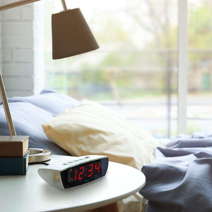 Lifestyle image of the 75908 0.9-inch Red LED Clear Case Alarm Clock shown on a night table next to a bed with large windows open onto a deck in the background. The digital alarm clock features red digits with a black lens, PM and ALARM indicators, and is in a clear case.