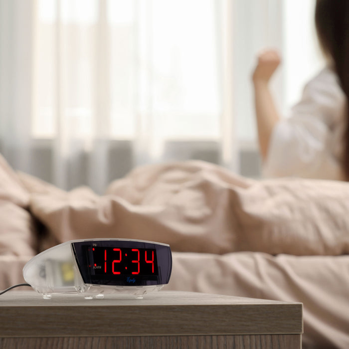 Lifestyle image of the 75908 0.9-inch Red LED Clear Case Alarm Clock shown on a night table next to a bed. The digital alarm clock features red digits with a black lens, PM and ALARM indicators, and is in a clear case.
