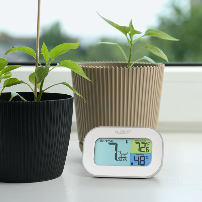 Image of the 919-147 Digital plant care timer with temperature and humidity. The timer sits on a windowsill with potted plants. Battery-operated, the timer features a white and gray case with a three section screen. The turquoise blue section displays how many days since the last watering (plant graphic with an up arrow) the yellow section lists the temperature at 72.6° F, and the dark blue section lists the humidity level at 48 percent with a water drop icon.