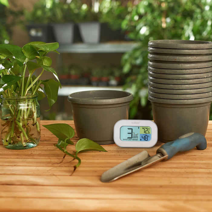Lifestyle image of the 919-147 Digital plant care timer with temperature and humidity. The timer is shown sitting on a potting bench with a stack of empty plant pots, plant clippings in a jar of water, and a planting trowel. The timer is battery-operated and features a white and gray oval case with a screen sectioned into three colors.