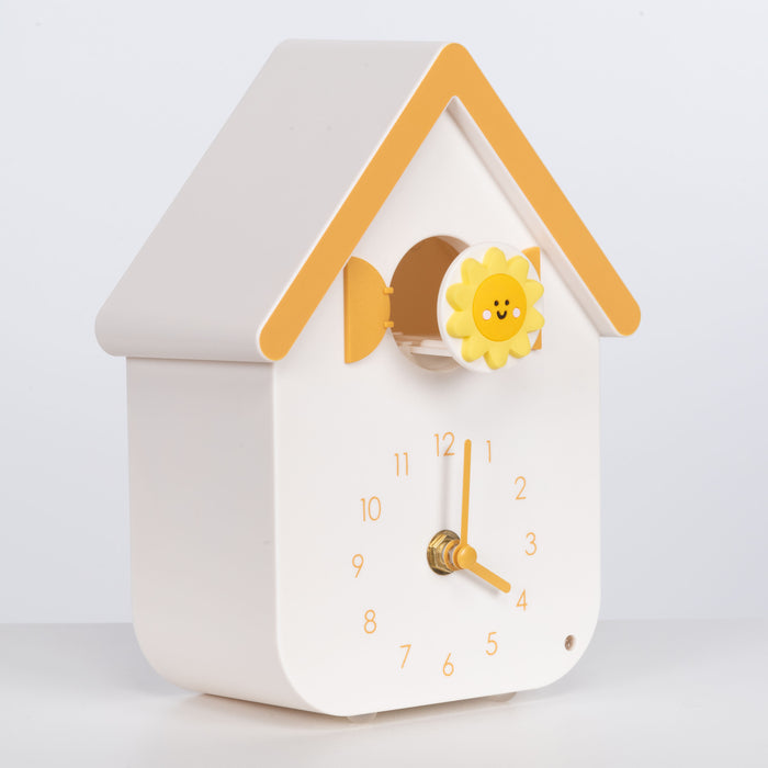 Right facing image of W70063, a white house shaped clock with a smiley sun cuckoo extended, with yellow numbers, shutters, roof, and hands