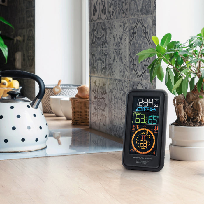 Lifestyle image of the S81120 Color Weather Station with Breeze shown sitting on a natural wood countertop next to a cook surface with a teapot. The color display features a vertical design with time, date, outdoor temperature, humidity, feels like reading, wind speed, and wind direction